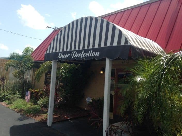 Exterior of Shear Perfection salon with striped awning and tropical landscaping.