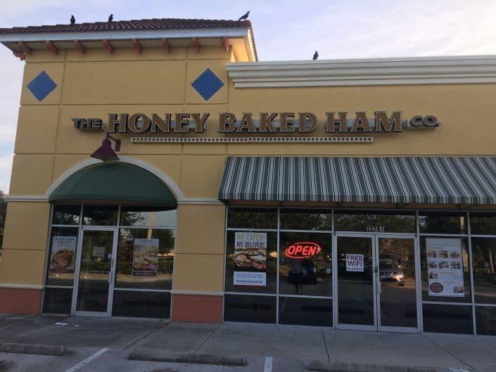 Exterior of The Honey Baked Ham Co. store with "Open" sign and shaded windows.